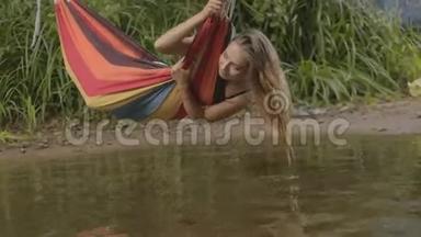 美丽的女孩坐在河边的吊床上，水触摸着手